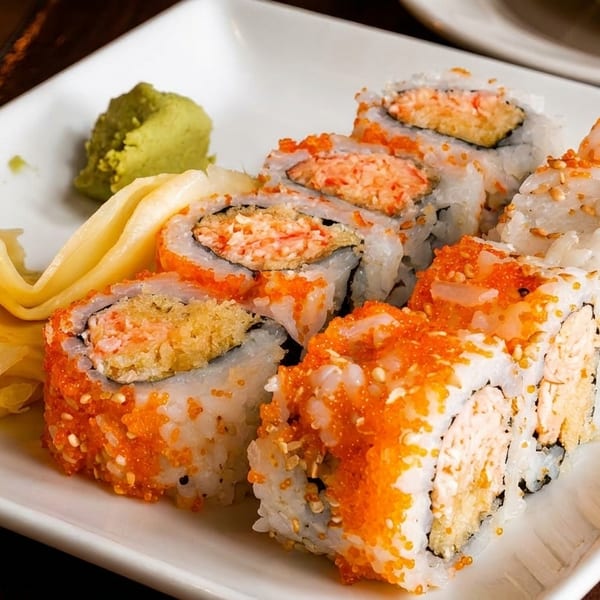 A white plate holds sushi rolls topped with orange roe, alongside pickled ginger and wasabi.