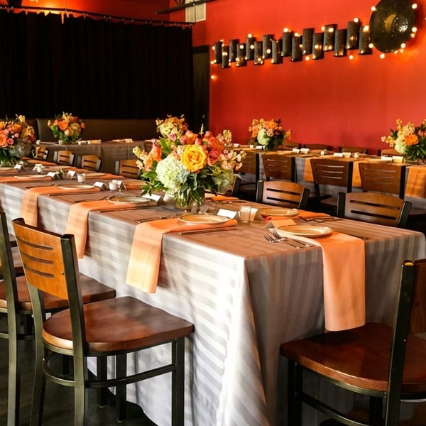 Long dining tables set with striped tablecloths, peach runners, and floral centerpieces in a restaurant.