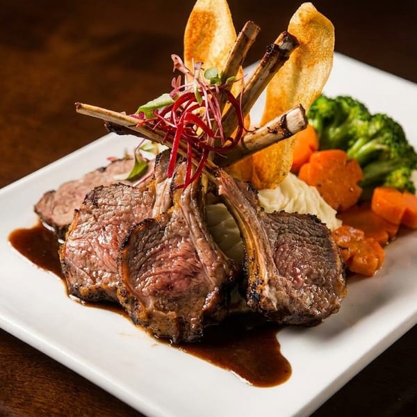 Rack of lamb with gravy, mashed potatoes, broccoli, carrots, and potato crisps on a white plate.