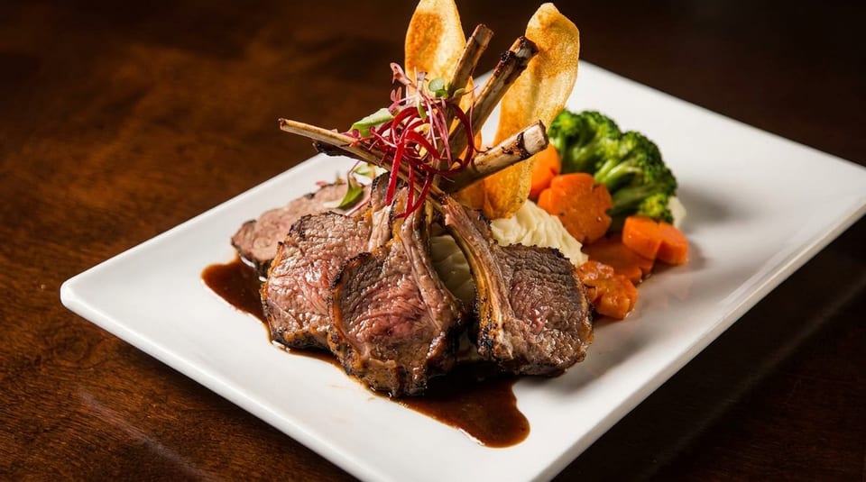 Plated rack of lamb with gravy, mashed potatoes, broccoli, carrots, and crispy potato slices.
