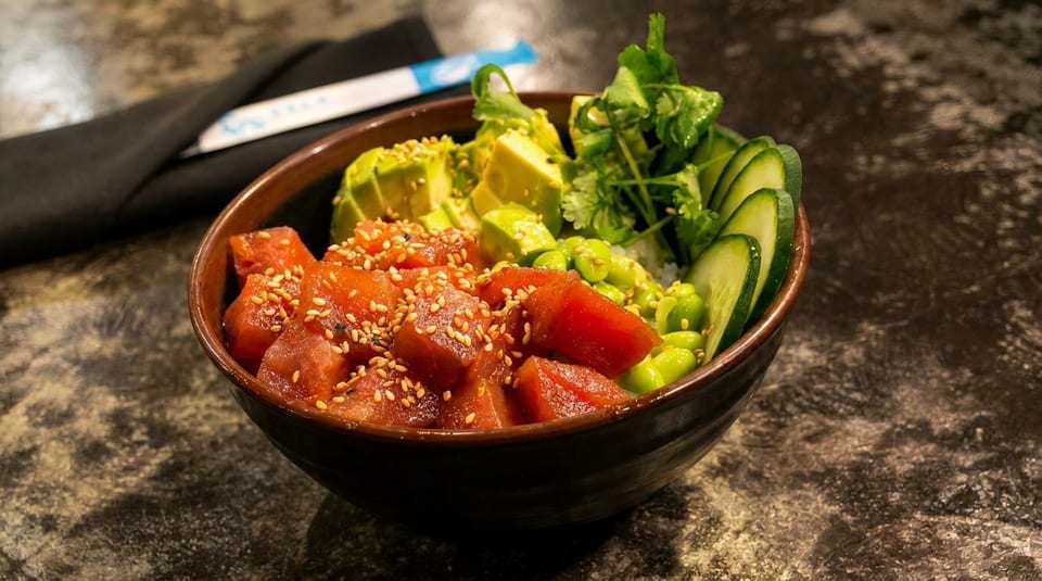 A vibrant poke bowl with tuna, avocado, edamame, cucumber, and cilantro on a dark table.
